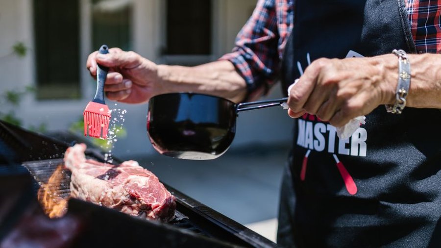 A person prepares a juicy steak on an outdoor grill, using a silicon brush to apply sauce.