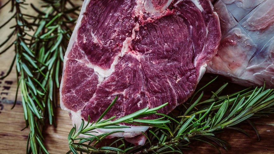 Bucată de carne crudă cu os, alături de crenguțe de rozmarin, pe un tocător de lemn