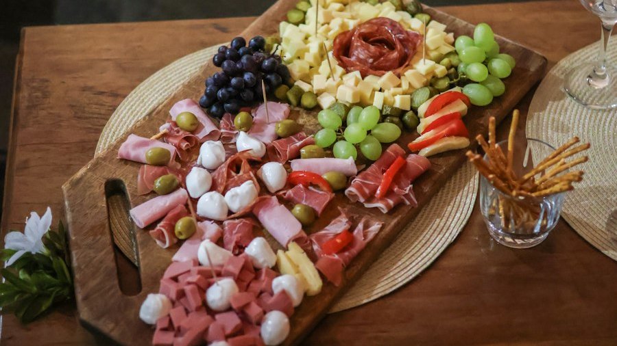 Platou de charcuterie pe tocător de lemn, cu mezeluri rulate, brânzeturi, struguri, măsline și grisine