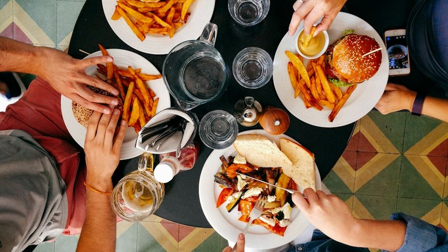 Mai multe persoane împart o masă la restaurant, cu burgeri, cartofi prăjiți și legume la cuptor